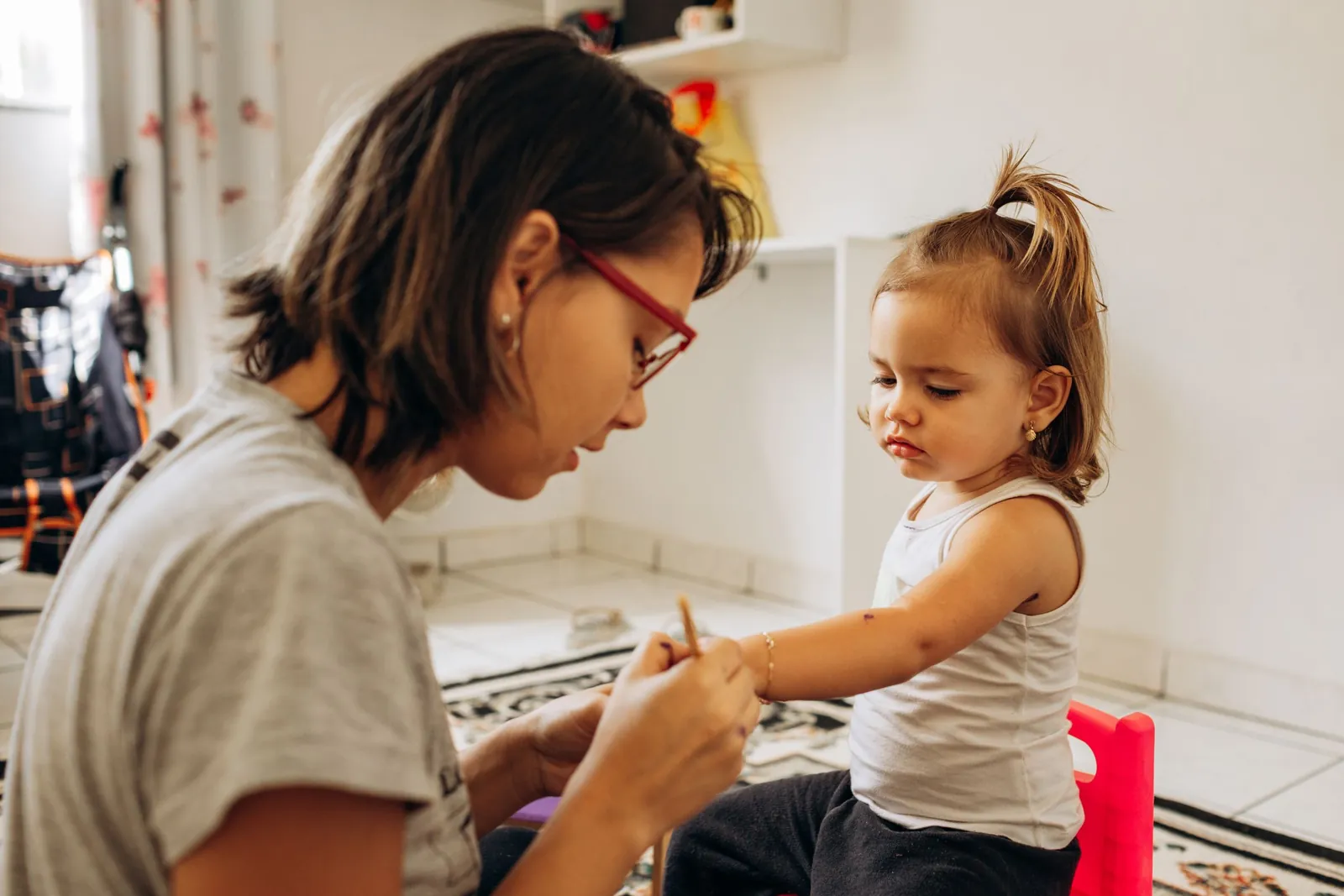 Una mamá con lentes guía a su hija pequeña en una manualidad, sentadas en el suelo de la casa