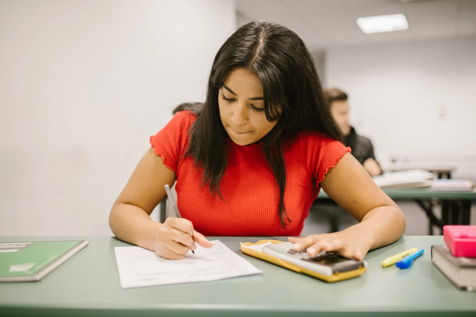 Estudiante escribe sus respuestas en una prueba, con una calculadora sobre la mesa