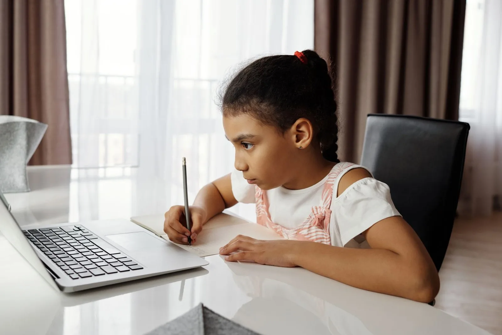 Niña escribe concentrada en su cuaderno, sentada frente a su computador portátil