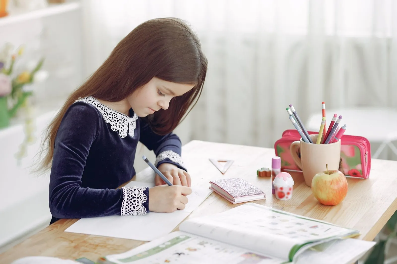 Niña escribe concentrada su tarea en la mesa de su casa, junto a sus lápices de colores y una manzana