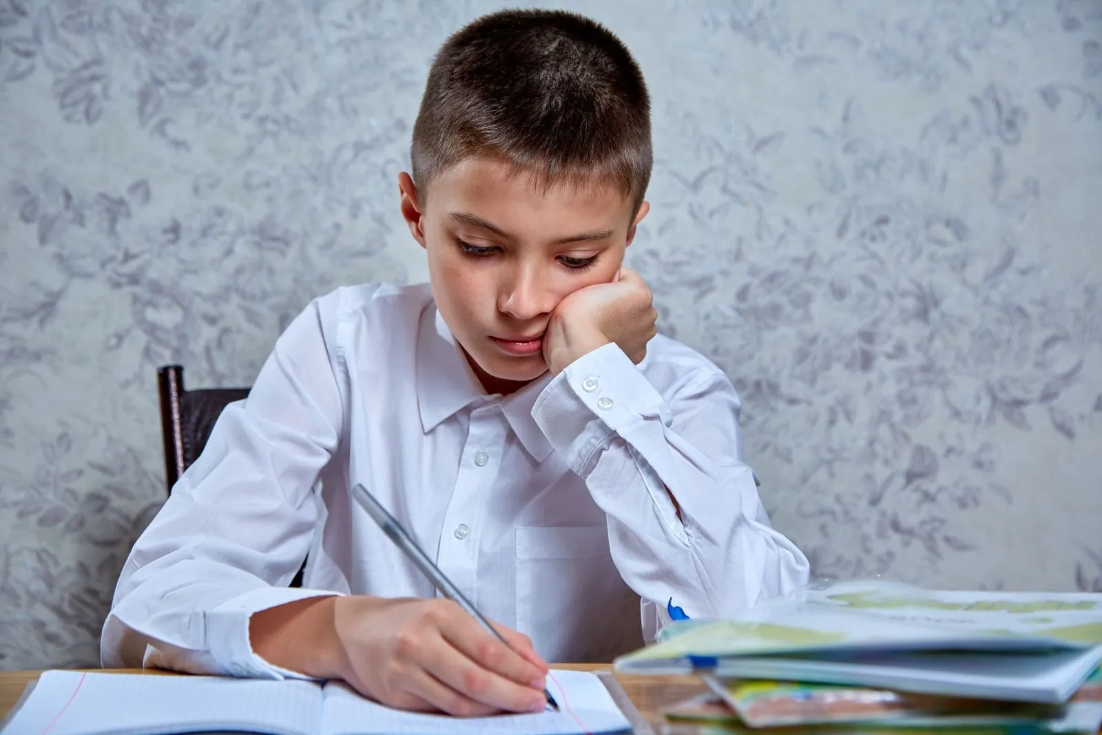 Niño con camisa blanca apoya la cara en la mano mientras escribe su tarea, cansado