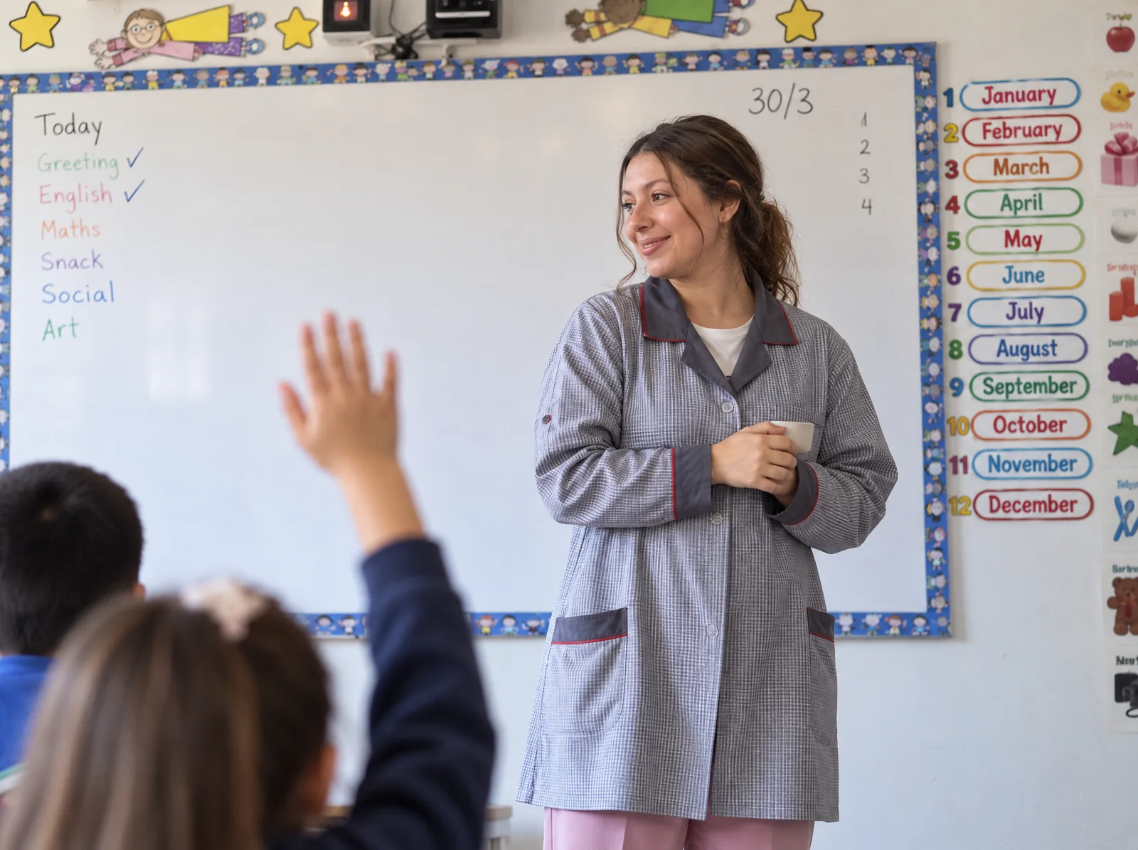 Isidora Benito frente a la pizarra de una sala de básica, junto a la lista de los meses en inglés, mientras una alumna levanta la mano