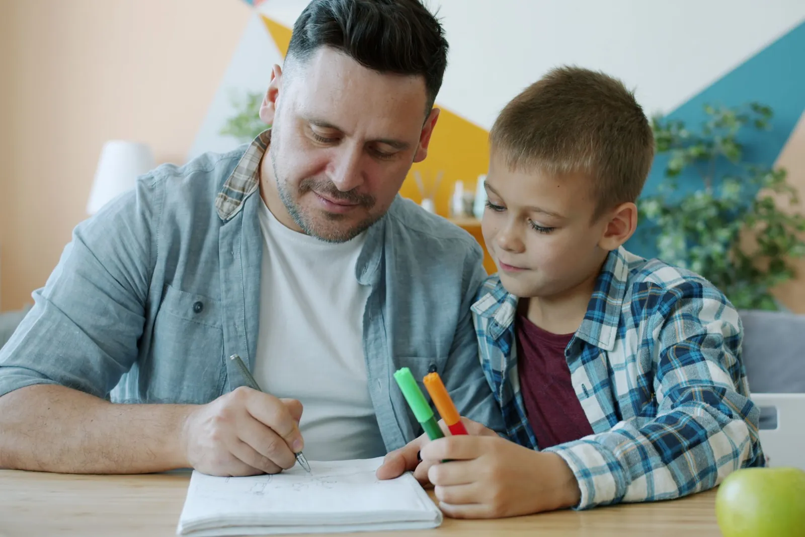 Un papá y su hijo hacen juntos una tarea en la mesa de la casa, con plumones de colores